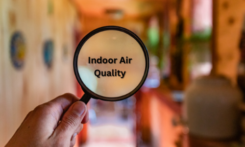 A person holding a magnifying glass with the text "Indoor Air Quality" written on it, focusing on the indoor environment. The background shows a blurred view of a room with warm lighting.