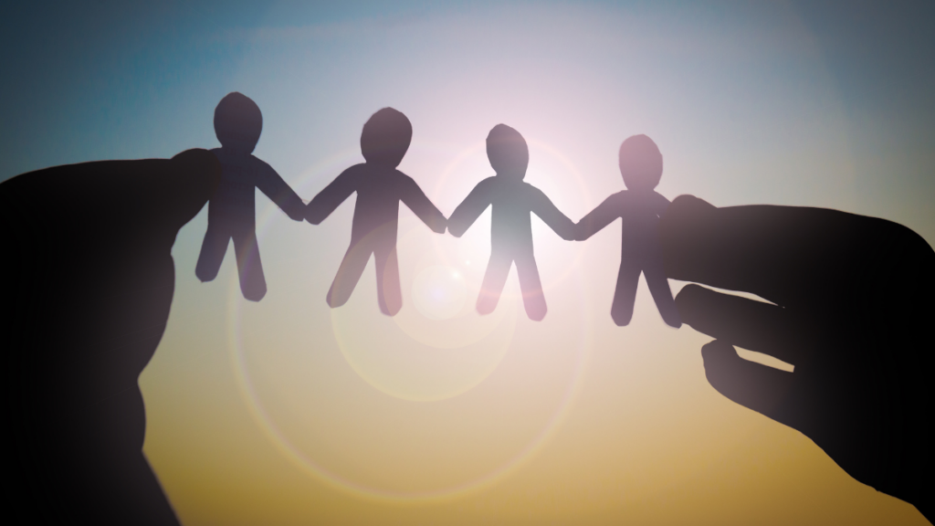 Silhouette of hands holding paper cutout figures of people in a chain, symbolizing unity and community. The image is backlit with a bright sunburst, creating a warm and hopeful atmosphere.