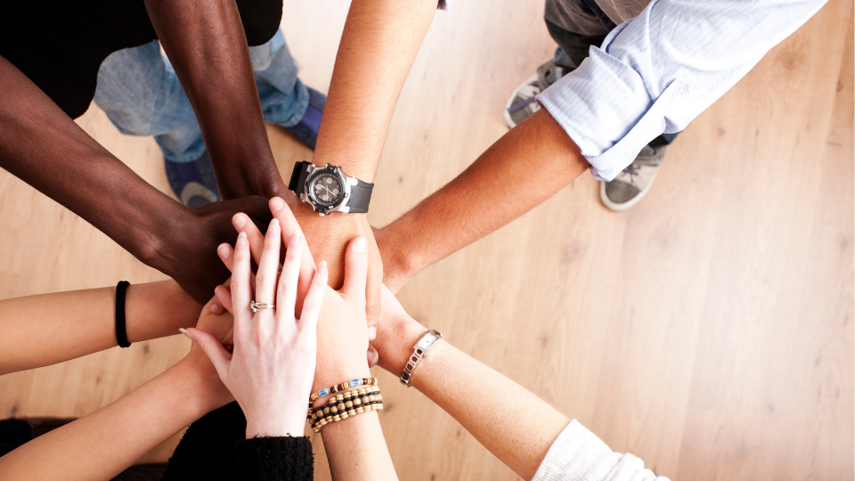 The image shows a group of diverse hands coming together in the center, symbolizing unity, teamwork, and collaboration. Each person has their hand stacked on top of the others, displaying a variety of skin tones, wrist accessories, and clothing styles, all emphasizing inclusivity and partnership. The background is a light wood floor.